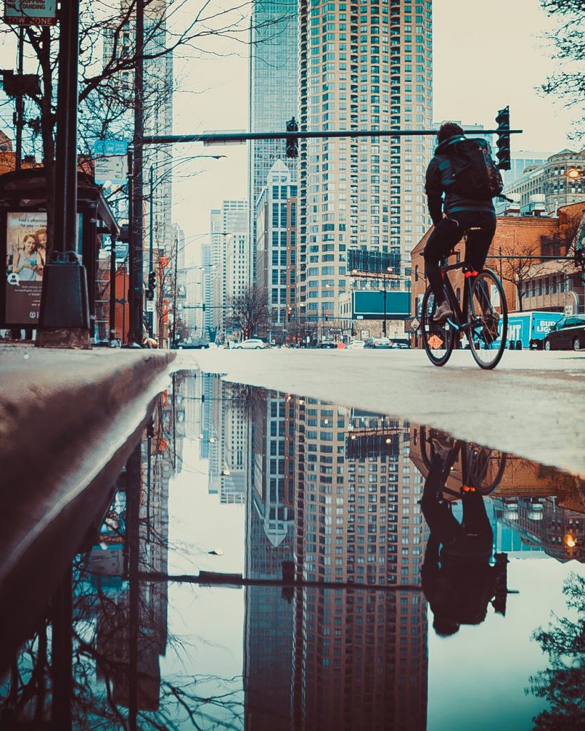 Ground-level view of a city street, with a large reflective puddle in the gutter and a person on a bicycle heading toward the large buildings of the downtown area.
